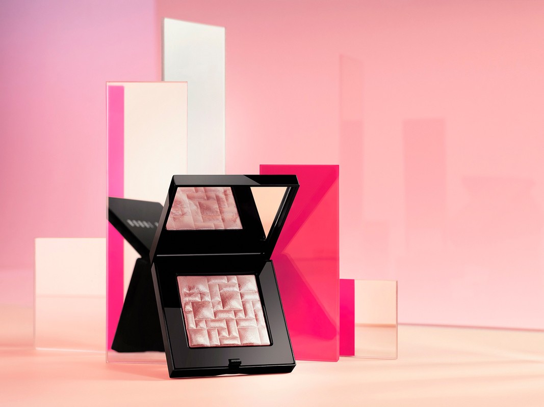 Still life studio shot of Bobbi Brown highlighting powder on a glossy light pink surface with translucent geometric block elements.