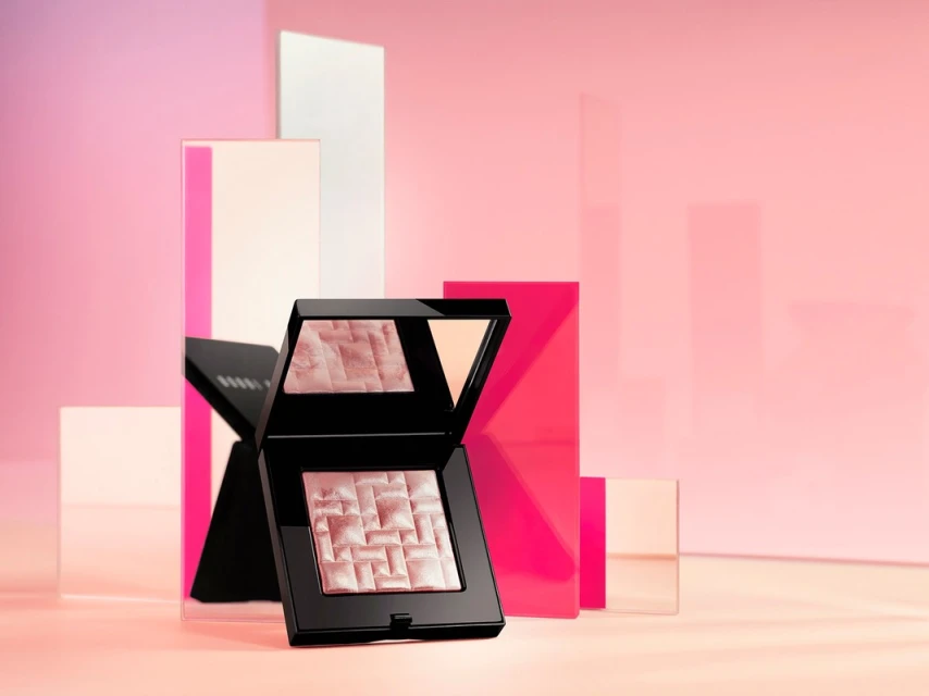 Still life studio shot of Bobbi Brown highlighting powder on a glossy light pink surface with translucent geometric block elements.
