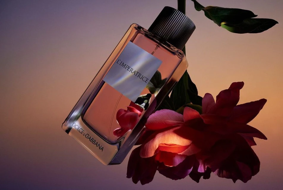Dolce & Gabbana L'Imperatrice perfume bottle with pink liquid, surrounded by pink peony flowers and green leaves on a diffused gradient background.