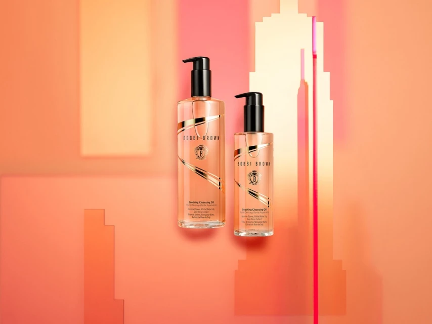 Two bottles of Bobbi Brown Soothing Cleansing Oil are displayed against a colorful gradient background in a beauty product shot.