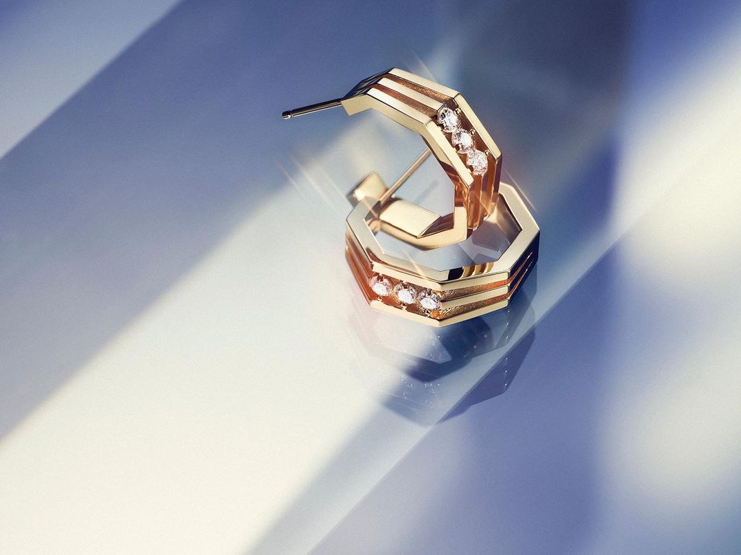 Pair of gold-tone hoop earrings with diamond accents on a reflective gradient blue surface with shadow and light play, product shot.