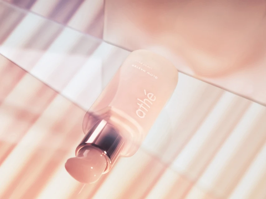 Close-up of a frosted pink 'athé Glow Master Vegan Collagen Boosting Serum' bottle with silver pump on a light pink striped surface.