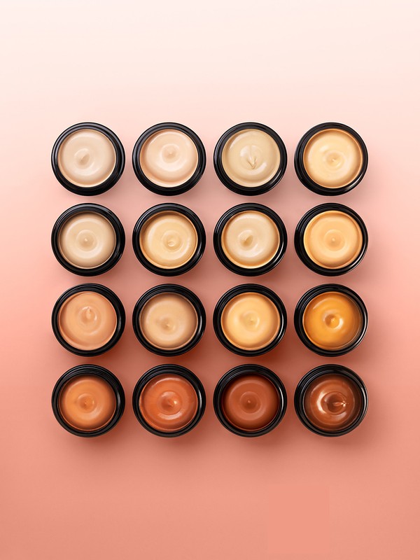 Overhead shot of sixteen containers with shades of cosmetic foundation, ranging from light to dark, on a peach background.