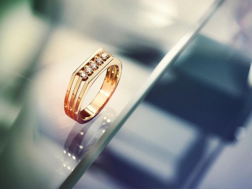Elegant gold ring featuring five small diamonds arranged in a row on a reflective glass surface, capturing a sense of luxury and craftsmanship.