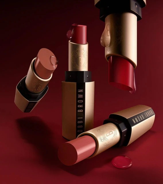 Studio shot of multiple Bobbi Brown lipsticks in varying red and nude shades against a dark red background, showcasing beauty products.