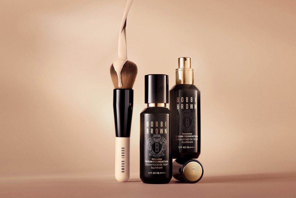 Cosmetics still life featuring Bobbi Brown serum foundation bottles, brush with foundation liquid on bristles, on a neutral background.