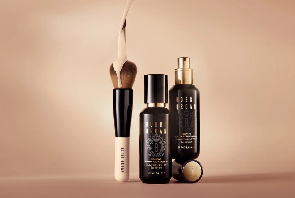 Cosmetics still life featuring Bobbi Brown serum foundation bottles, brush with foundation liquid on bristles, on a neutral background.