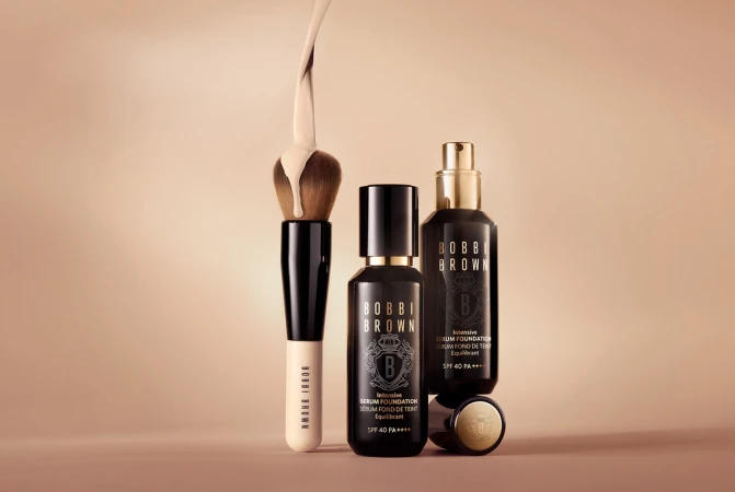 Cosmetics still life featuring Bobbi Brown serum foundation bottles, brush with foundation liquid on bristles, on a neutral background.