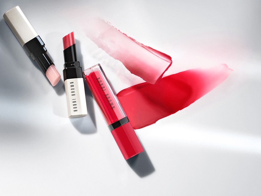 Cosmetic still life of Bobbi Brown lipsticks in red and pink shades, along with lipstick smears, set against a white backdrop.