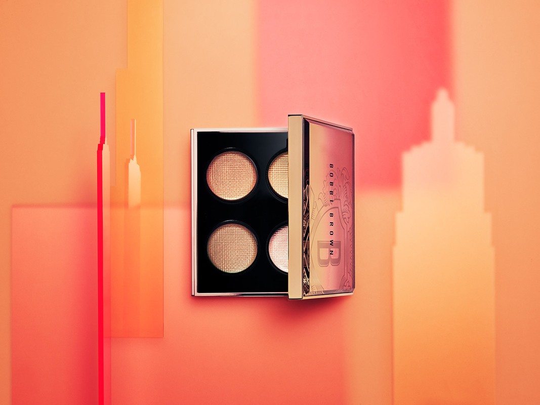 Bobbi Brown eyeshadow palette featuring four shades of shimmering golden and bronze tones, presented on a peach-colored backdrop with geometric shapes.