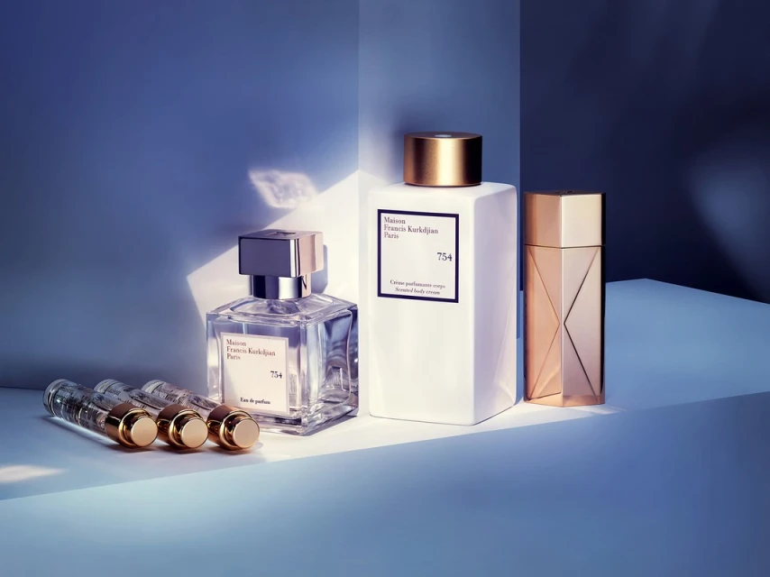Four perfume bottles, three of which are miniature, stand on a white reflective surface with soft blue lighting behind them.