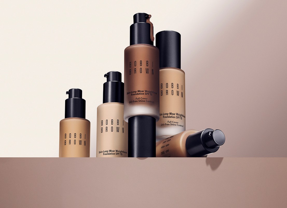 Bobbi Brown foundation bottles of various shades are arranged on a beige surface. The product is in focus and well-lit.