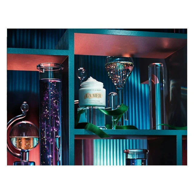 Still life of La Mer moisturizing cream with clear glasses and green ribbon on a teal shelf against a backdrop of vertical teal lines.
