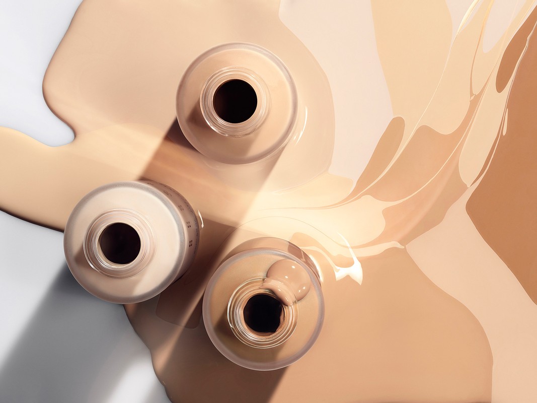 Overhead studio shot of three foundation bottles with various shades of tan foundation fluid spilled around the product.