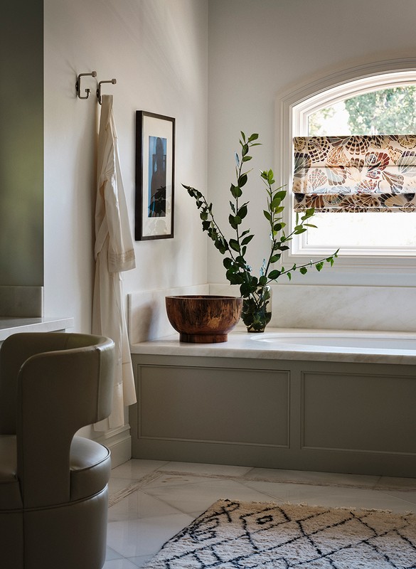 Modern bathroom interior featuring a freestanding tub, patterned window shade, wooden bowl, vase with greenery, and a unique chair.