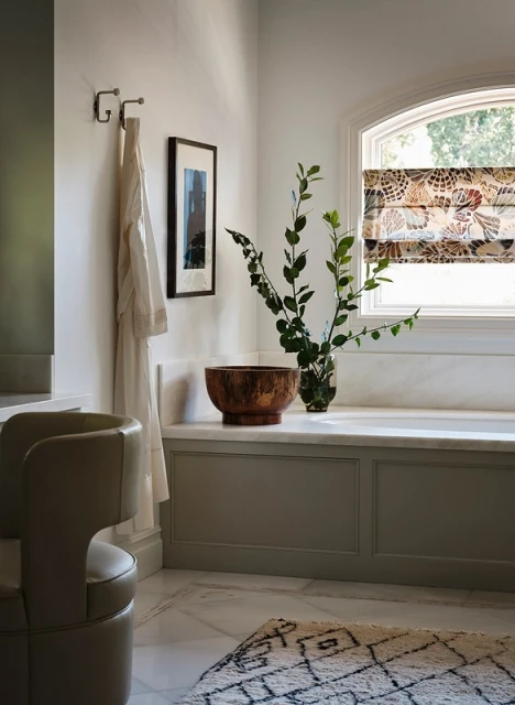 Modern bathroom interior featuring a freestanding tub, patterned window shade, wooden bowl, vase with greenery, and a unique chair.