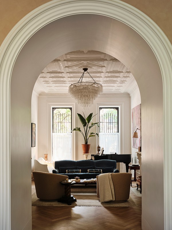 Living room interior featuring a blue velvet sofa, two beige armchairs, a black piano, a decorative chandelier, and natural light from windows.
