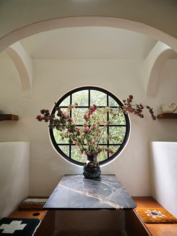 Interior dining area featuring a marble table with floral arrangement, round window, white walls, arched doorways, and built-in bench seating.