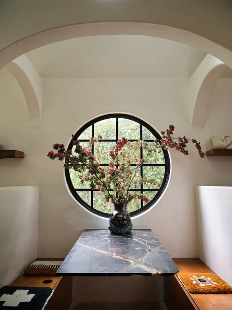 Interior dining area featuring a marble table with floral arrangement, round window, white walls, arched doorways, and built-in bench seating.