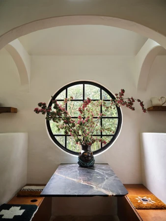 Interior dining area featuring a marble table with floral arrangement, round window, white walls, arched doorways, and built-in bench seating.