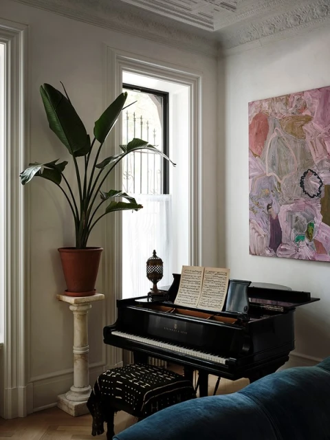 Interior corner featuring a black grand piano with sheet music, a potted plant on a pedestal, abstract art, and a patterned stool.