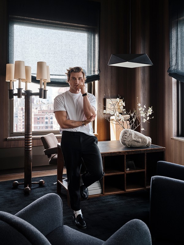 Indoor portrait of a fair-skinned adult male with blond hair wearing casual clothes, standing near a desk and large lamp in a modern room.