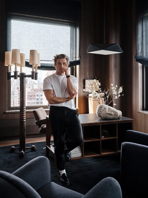 Indoor portrait of a fair-skinned adult male with blond hair wearing casual clothes, standing near a desk and large lamp in a modern room.