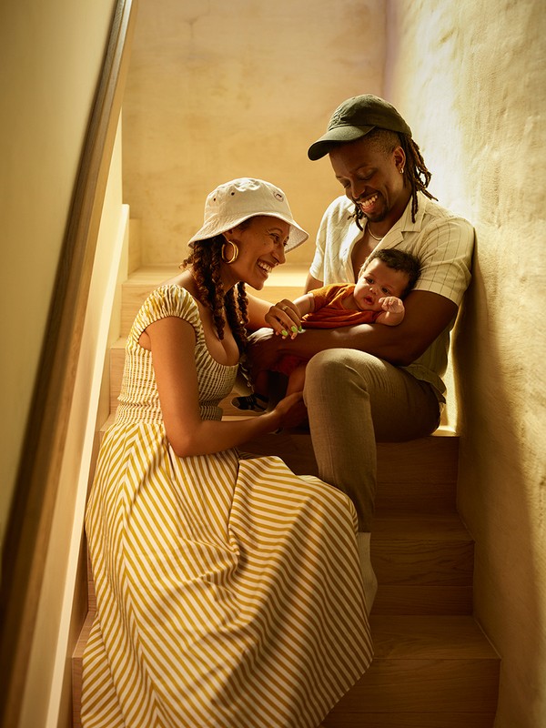 Family portrait on stairs: parents smiling at their baby. Mother in a striped dress, father in a cap, capturing a tender family moment.