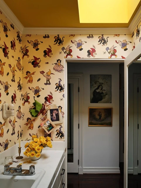 Whimsical bathroom interior featuring cartoon character wallpaper, a white countertop with yellow flowers, and artwork in a hallway.