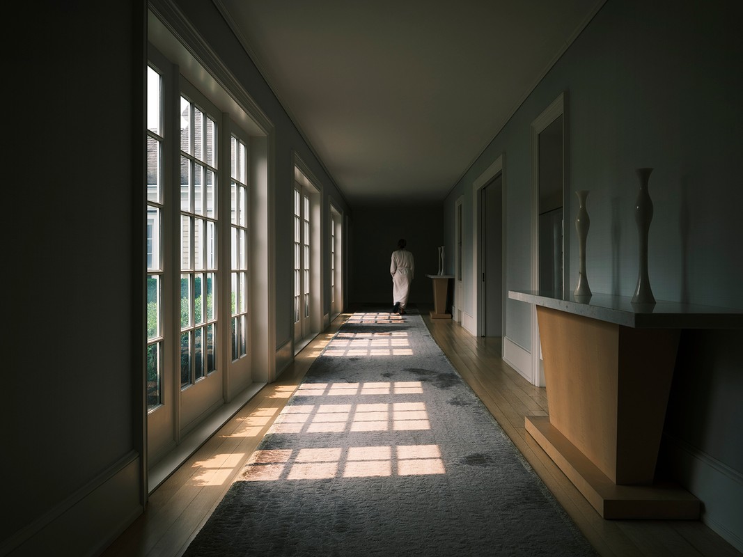 Woman in white robe walking down a long hallway with windows, natural light casting shadows on the floor. Interior design elements.
