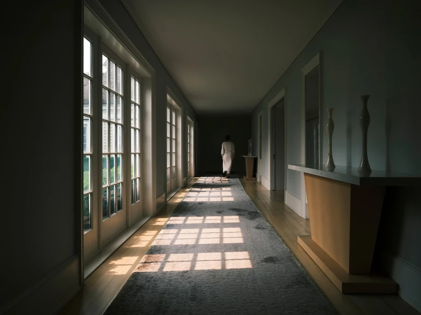Woman in white robe walking down a long hallway with windows, natural light casting shadows on the floor. Interior design elements.