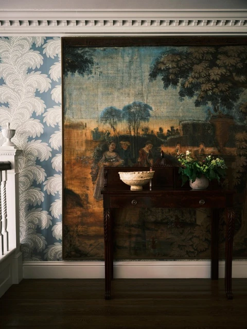 Interior view with a dark wood table supporting a white bowl and flowers, set against a tapestry and floral wallpaper background.
