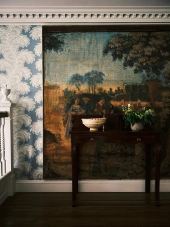 Interior view with a dark wood table supporting a white bowl and flowers, set against a tapestry and floral wallpaper background.