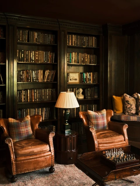 Cozy reading nook featuring two brown leather armchairs with plaid pillows, a chess set, and a bookshelf in a dimly lit room.