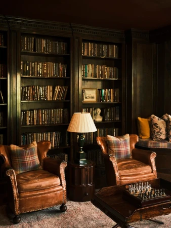Cozy reading nook featuring two brown leather armchairs with plaid pillows, a chess set, and a bookshelf in a dimly lit room.