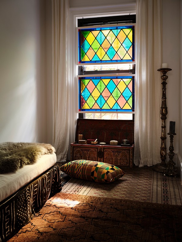 Bedroom interior featuring a bed with a patterned base, colorful pillows, a stained-glass window, and vintage wooden chest.