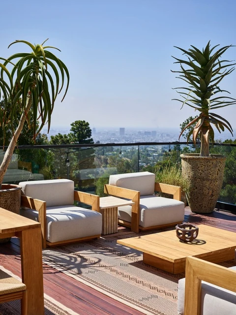 Outdoor patio featuring armchairs, a coffee table, and potted plants with a cityscape view on a sunny day.