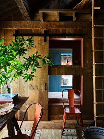 Rustic interior with wooden walls, barn door, orange metal chairs, ladder, and green foliage, showcasing a blend of textures and colors.