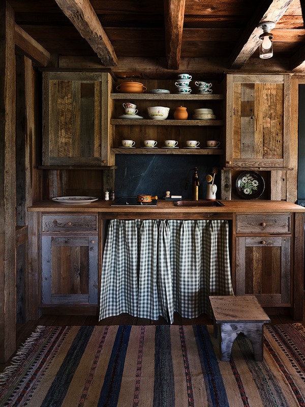 Rustic kitchen interior featuring wooden cabinets, shelves with dishes, a checkered curtain, and a striped rug, creating a warm, cozy atmosphere.