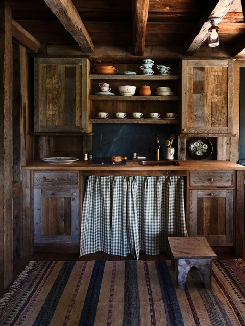 Rustic kitchen interior featuring wooden cabinets, shelves with dishes, a checkered curtain, and a striped rug, creating a warm, cozy atmosphere.