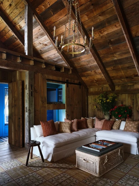 Rustic living room interior with white slipcovered sofa, wood-paneled walls and ceiling, wicker trunk, stool, and decorative chandelier.