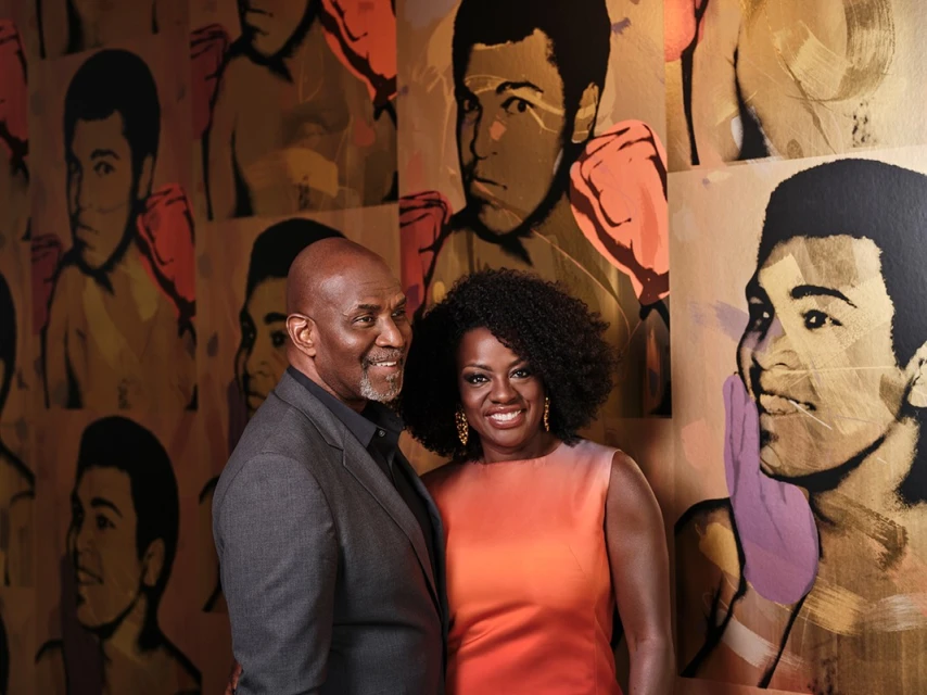Couple poses in front of colorful art featuring portraits of a boxer in varying artistic styles; the art covers the entire background.