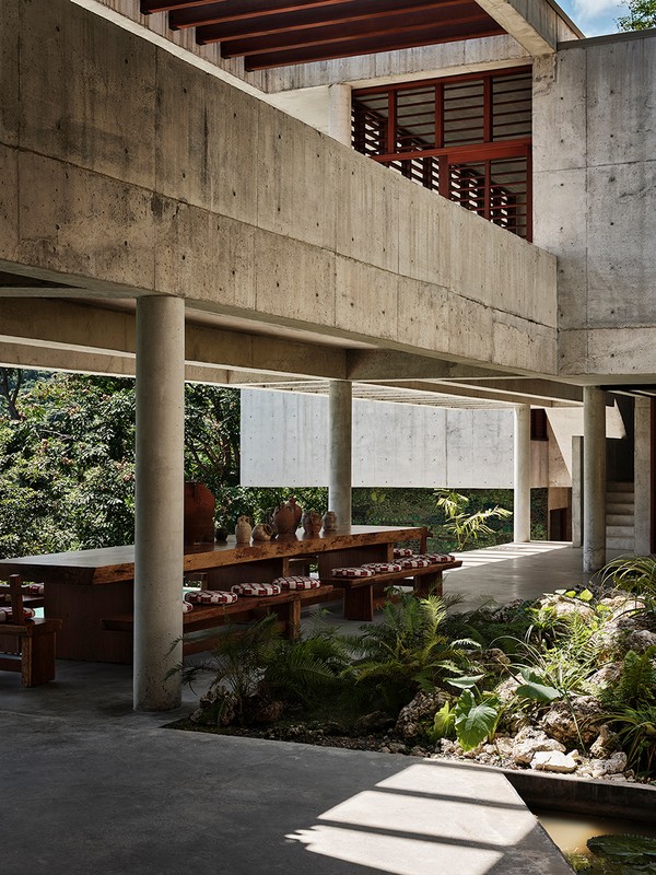 Exterior view of a modern concrete building with a long wooden table and benches, surrounded by lush greenery and plants.