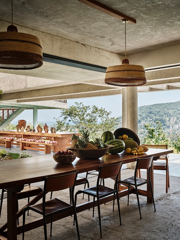 Indoor dining area features a long wooden table set with fresh fruit, flanked by black chairs, and lit by pendant lights.