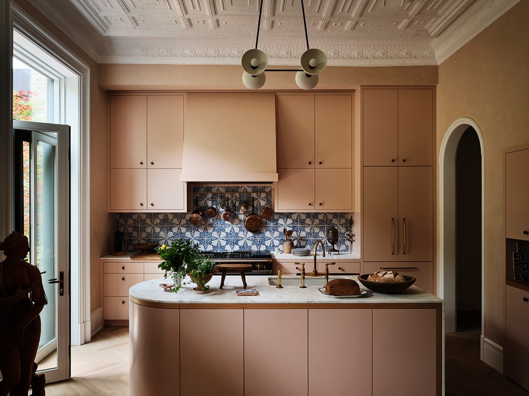 Elegant kitchen interior featuring blush pink cabinetry, marble island, patterned tile backsplash, and decorative ceiling in a brightly lit space.