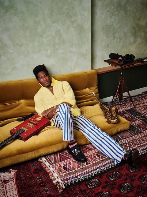 Stylish African American male musician poses on a gold velvet couch with a red guitar and saxophone in a naturally lit interior.