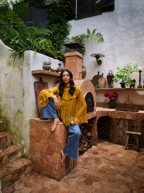 Adult female with bare feet sits on stone oven in rustic outdoor kitchen with tile floor, wearing a flowing gold top and wide leg jeans.