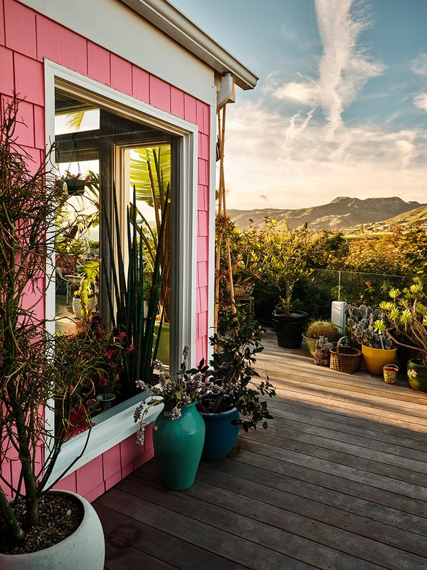 A pink house corner with a window is seen from a wooden deck. Various potted plants are placed on the deck and in the window box.