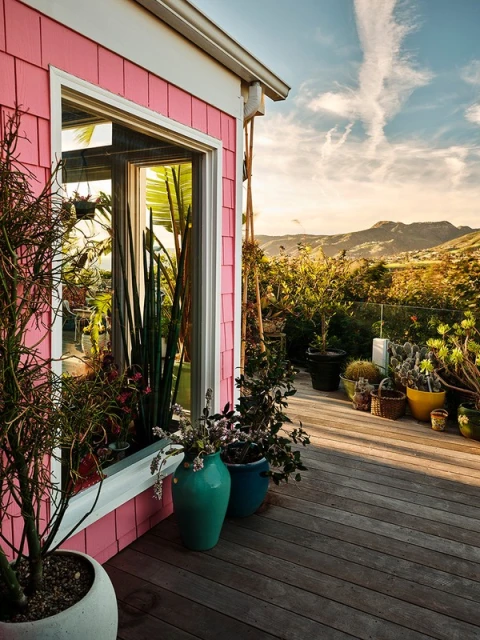A pink house corner with a window is seen from a wooden deck. Various potted plants are placed on the deck and in the window box.