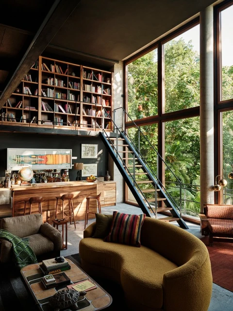 An interior living space showcasing a mustard-colored sofa, wooden bar with stools, and expansive windows revealing lush outdoor greenery.